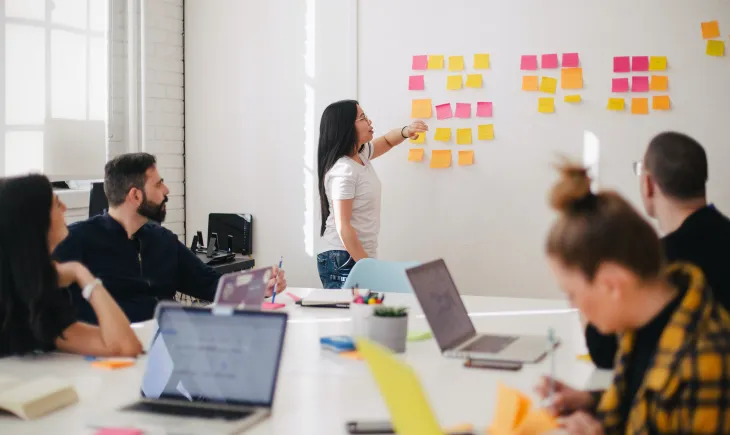 Woman presenting to table of people pointing at sticky notes on wall